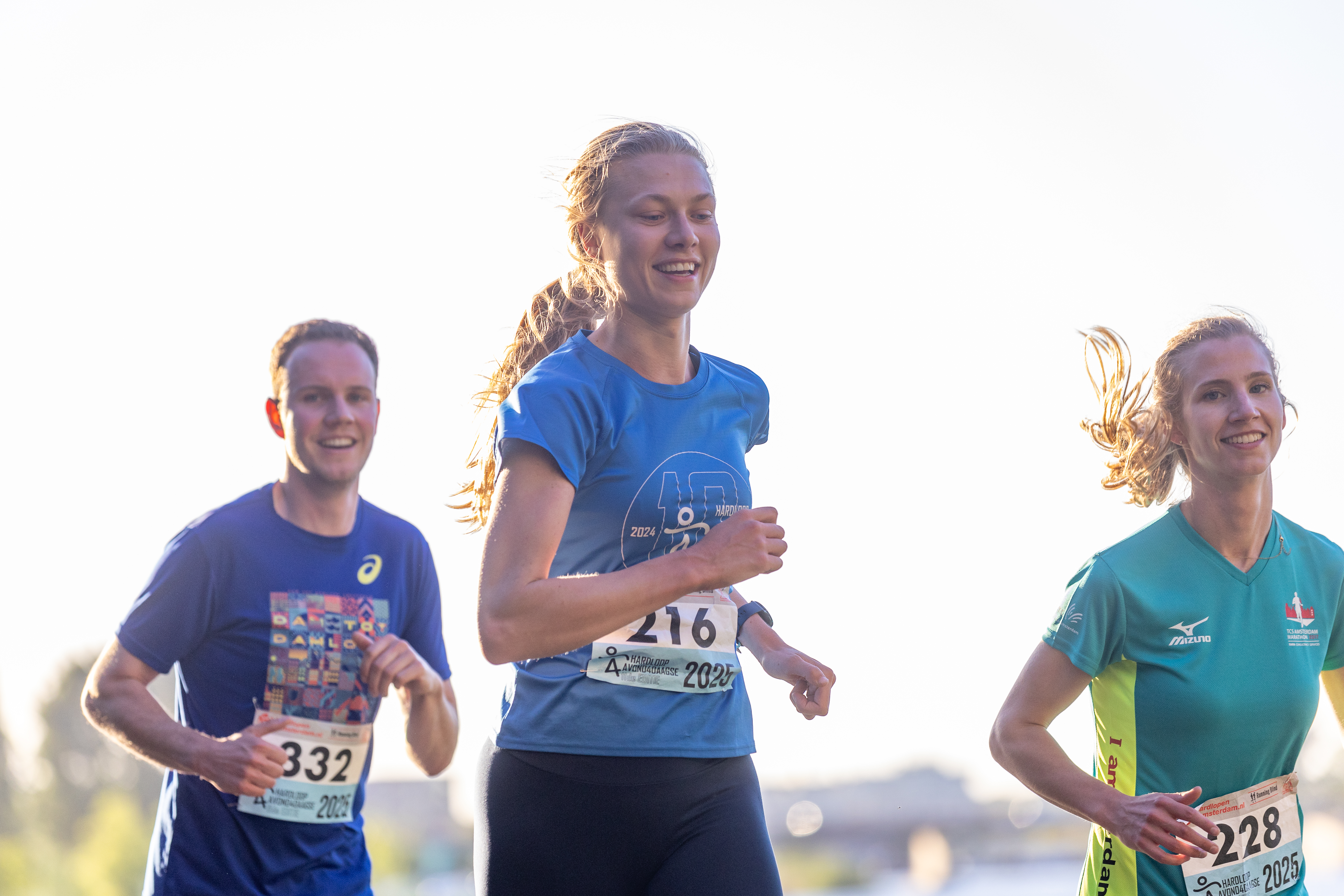 Drie hardlopers lopen in fel tegenlicht langs het water.