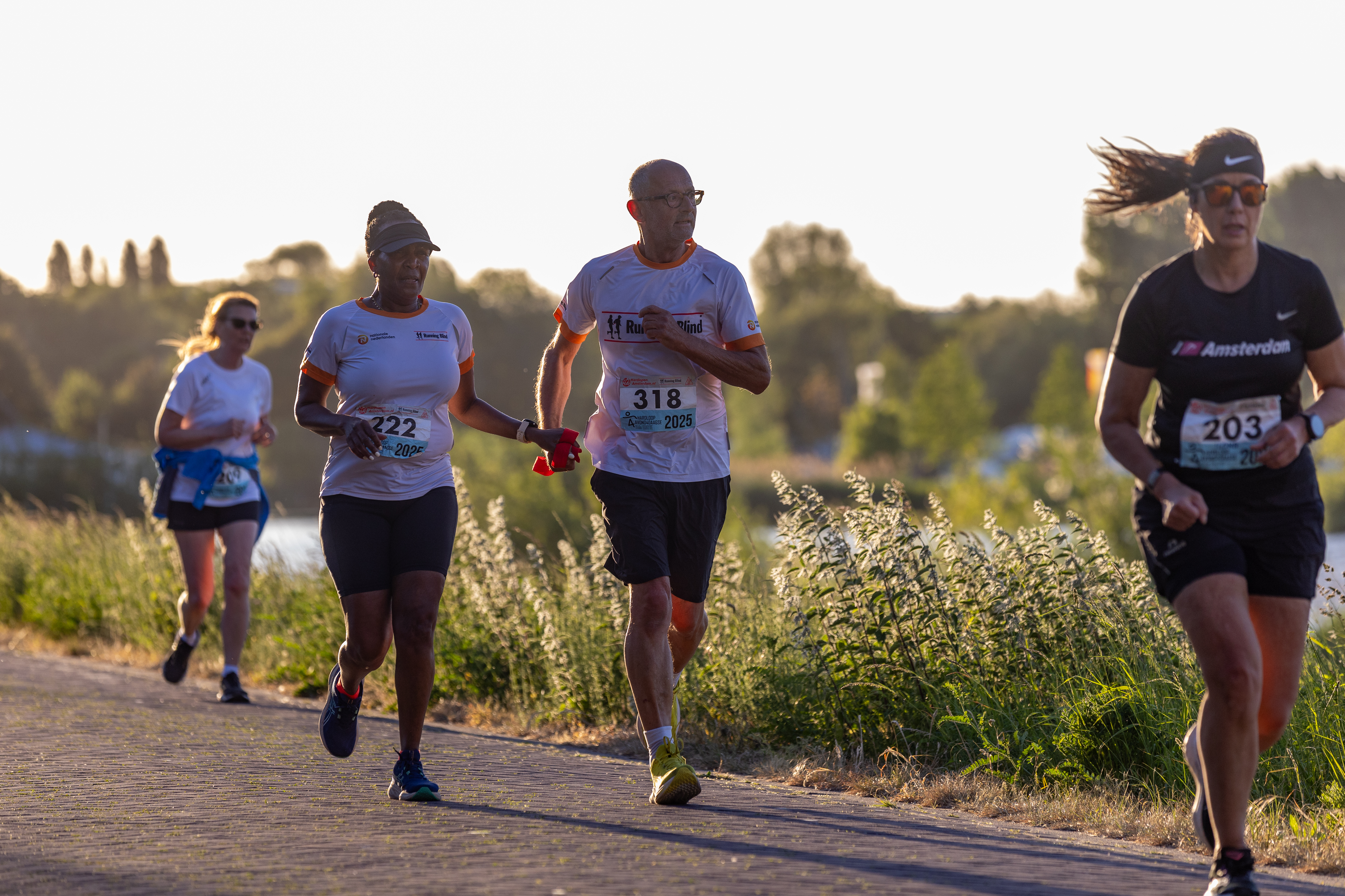 Deelnemers lopen in avondlicht langs het water, met drie lopers voorop.