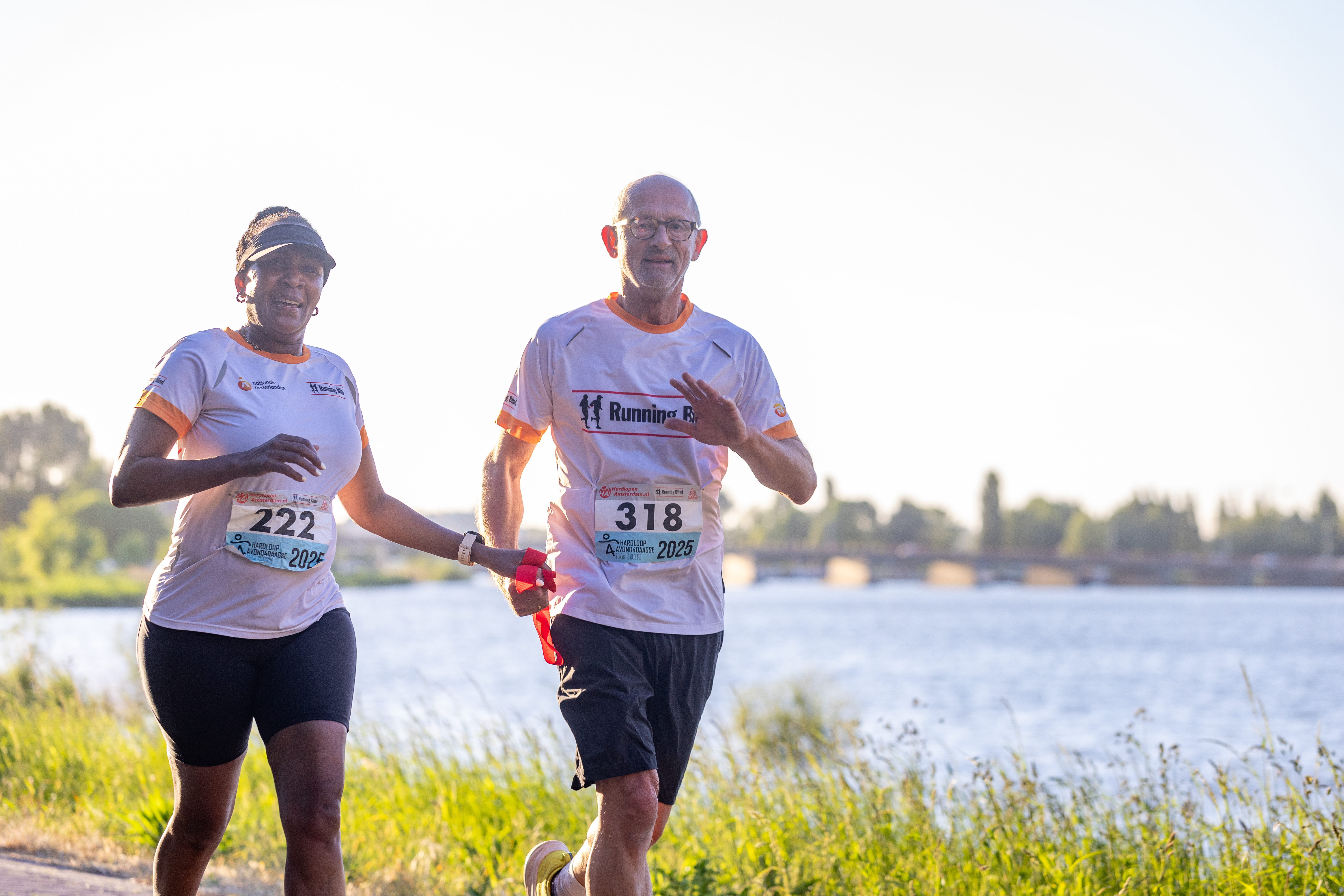 Een Running Blind-duo loopt met een rood verbindingslint langs het water.