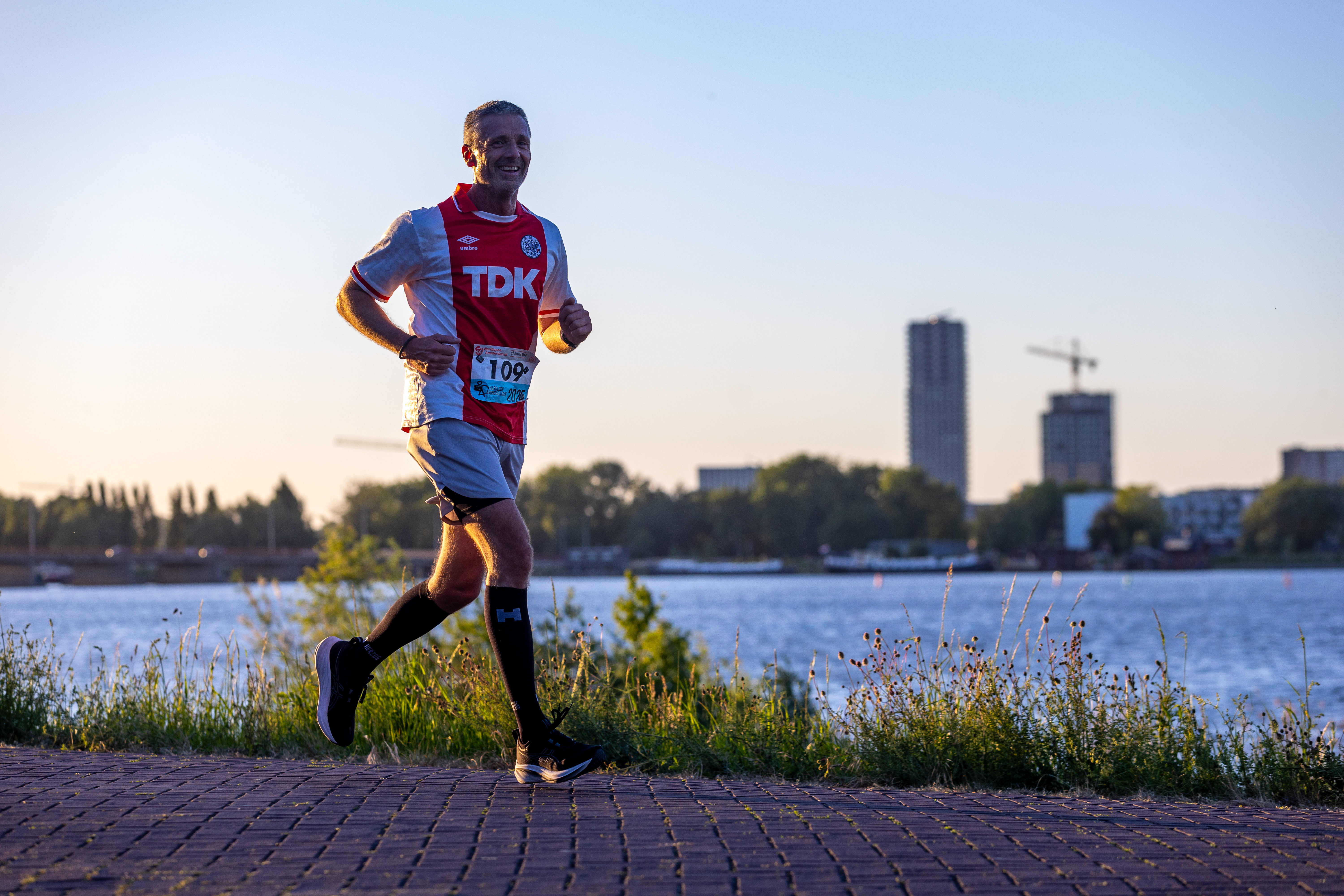Een hardloper in rood-blauw tenue loopt langs het water in de avondzon.