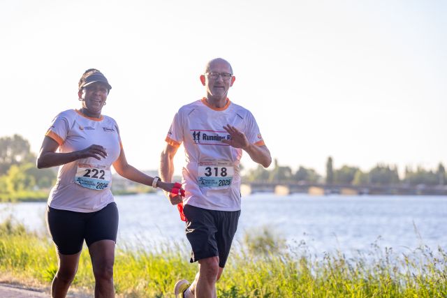 Een Running Blind-duo loopt met een rood verbindingslint langs het water.