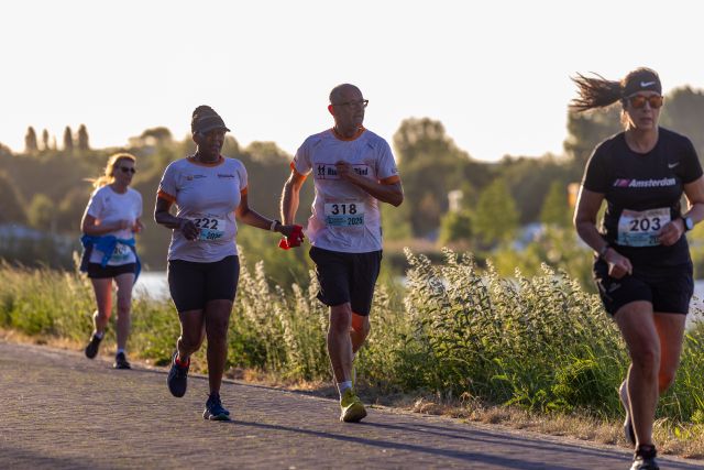 Deelnemers lopen in avondlicht langs het water, met drie lopers voorop.