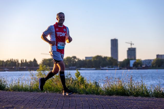 Een hardloper in rood-blauw tenue loopt langs het water in de avondzon.